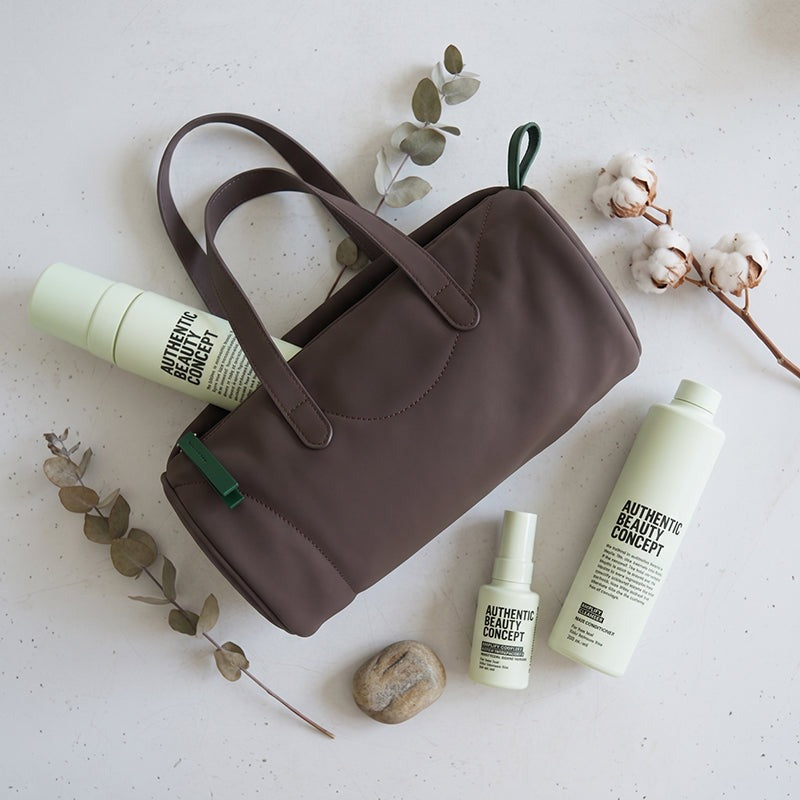 A brown nylon cosmetic bag with dual top handles is arranged on a light speckled surface with botanical props and amplify products from Authentic Beauty Concept.
