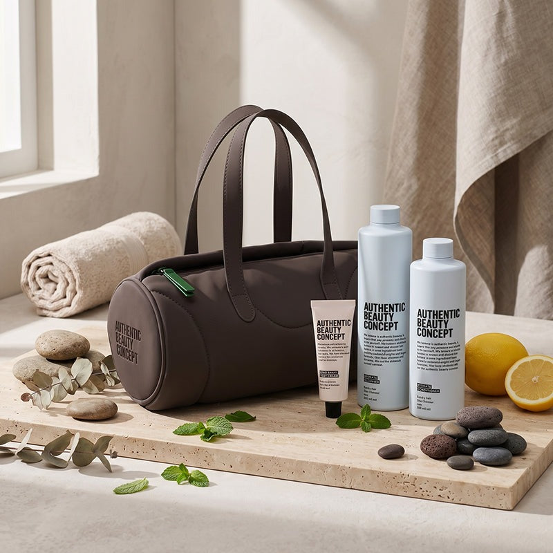 A dark brown cosmetic tote bag with 'AUTHENTIC BEAUTY CONCEPT' branding sits on a wooden board next to hydrate spire bottles and hand cream, styled with spa props like stones, mint leaves, a lemon, and a rolled towel.