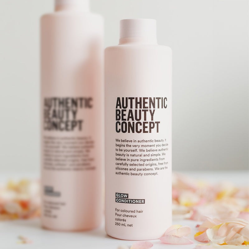 A bottle of Authentic Beauty Concept Glow Conditioner in the foreground, with a second matching bottle blurred in the background. The bottles are a very pale pinkish-white with black text, set against a clean white background with soft pink flower petals scattered at the base.