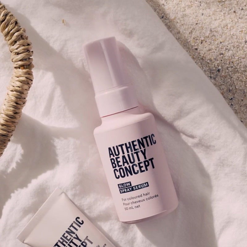 Glow spray serum bottle labeled 'AUTHENTIC BEAUTY CONCEPT' and a matching cream tube resting on a white textured fabric beside a woven straw accessory, in warm natural light.