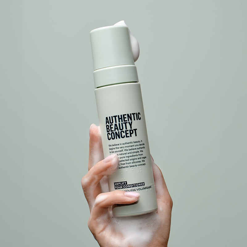 A hand holding a frosted white pump bottle of 'Authentic Beauty Concept' serum against a solid light gray background, with a dollop of white serum visible at the nozzle.