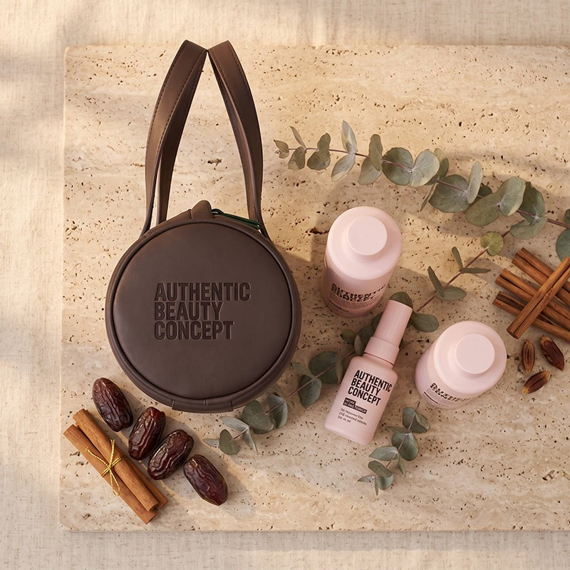A round dark brown Authentic Beauty Concept makeup bag with dual shoulder straps, styled in a flat lay on a beige stone surface with Glow products, eucalyptus leaves, cinnamon sticks, and dates.