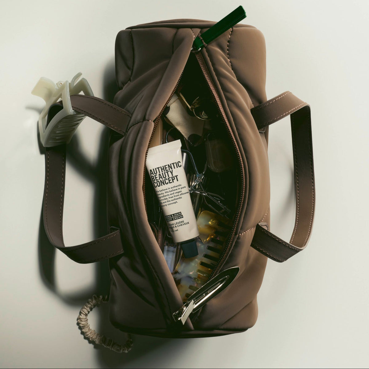 A brown nylon cosmetic bag with its zipper open, styled with various beauty products inside, shot from a top-down perspective on a light gray background.