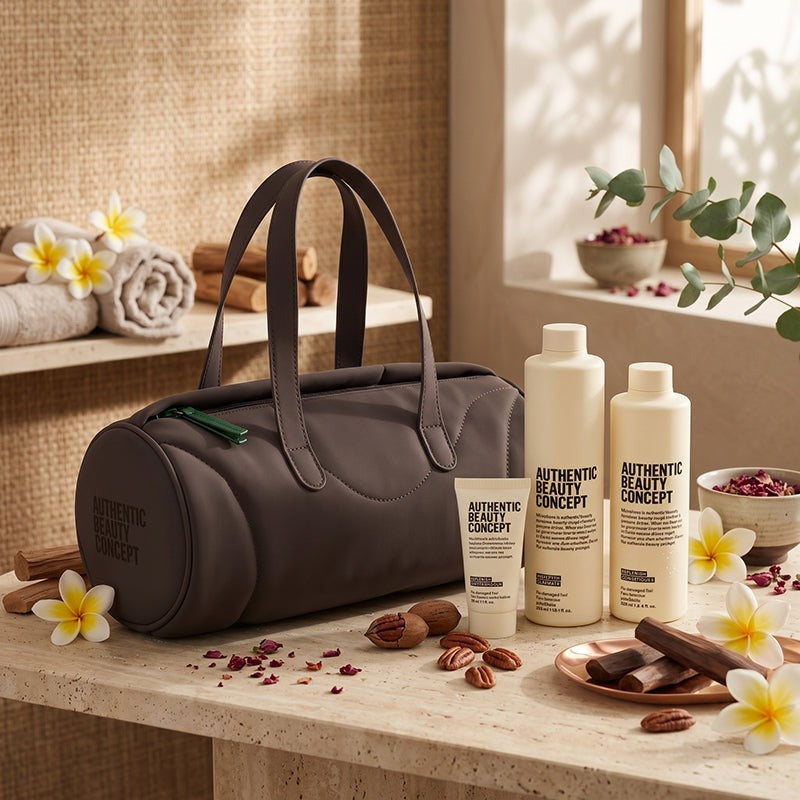 A dark brown barrel cosmetic bag with two handles sits on a stone surface beside three Replenish bottles and tubes labeled 'AUTHENTIC BEAUTY CONCEPT.' The scene is styled with towels, flowers, nuts, dried petals, and greenery in a warm spa-like interior.
