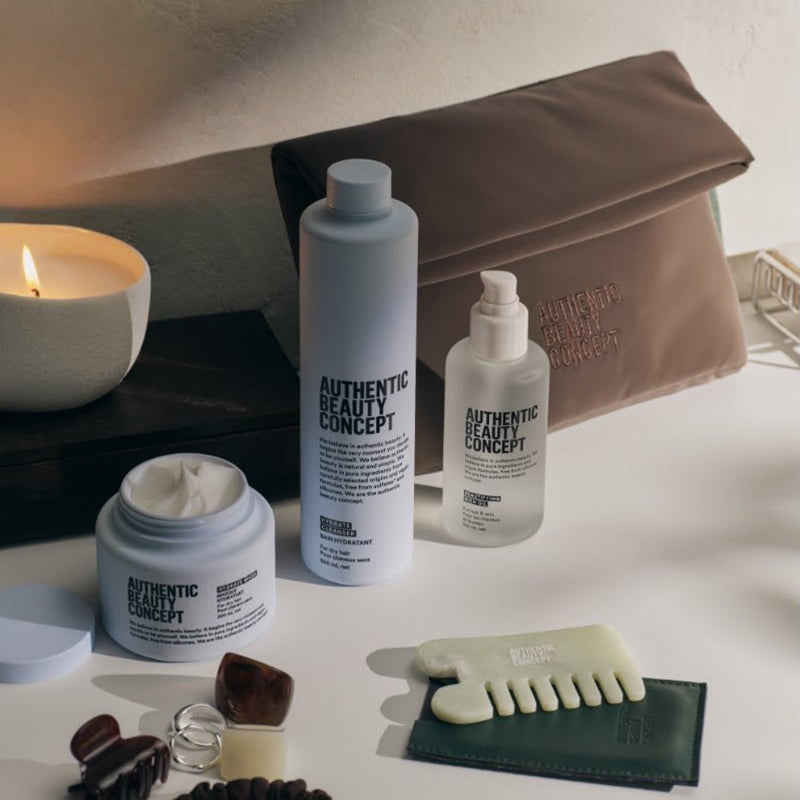 A curated beauty product arrangement on a tabletop featuring hydrate cleanser, hair oil, a hydrate mask, a brown pouch with gold text, a green comb and pouch, and small accessories, with a lit candle in the background.