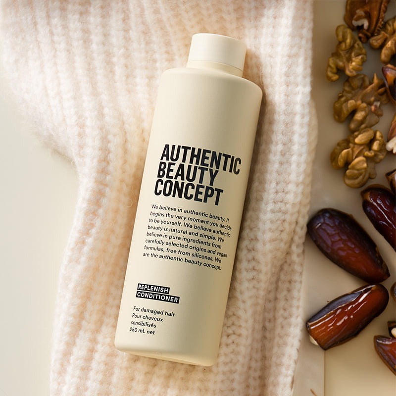 A bottle of Authentic Beauty Concept Replenish Conditioner lies on a textured cream knit blanket, surrounded by natural ingredients like dates and walnuts on a light beige surface.