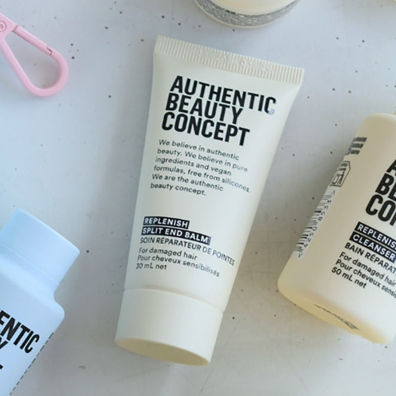A white tube of Authentic Beauty Concept Replenish Split End Balm is shown on a light speckled surface, with partial views of other skincare bottles nearby.