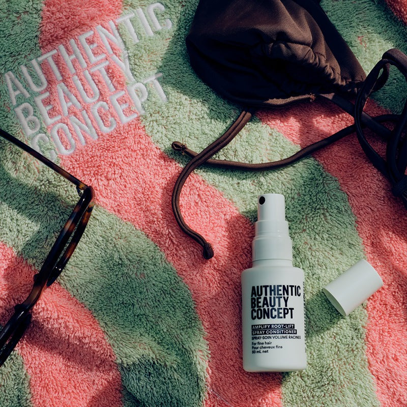 Amplify root-lift spray conditioner labeled 'AUTHENTIC BEAUTY CONCEPT' lies on a pink and green patterned towel, with a brown drawstring bag, eyeglasses, and a small white cap nearby. The words 'AUTHENTIC BEAUTY CONCEPT' are also printed on the towel.