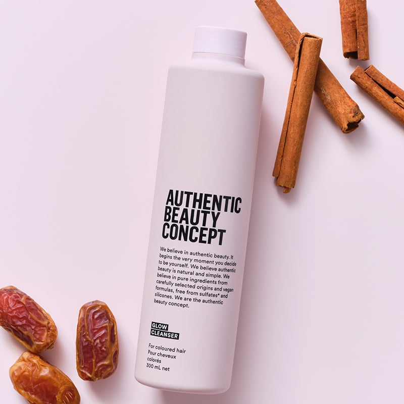 A 300 mml glow cleanser of 'Authentic Beauty Concept' Skin Cleanser, styled in a flat-lay with cinnamon sticks and dates on a light pink background.