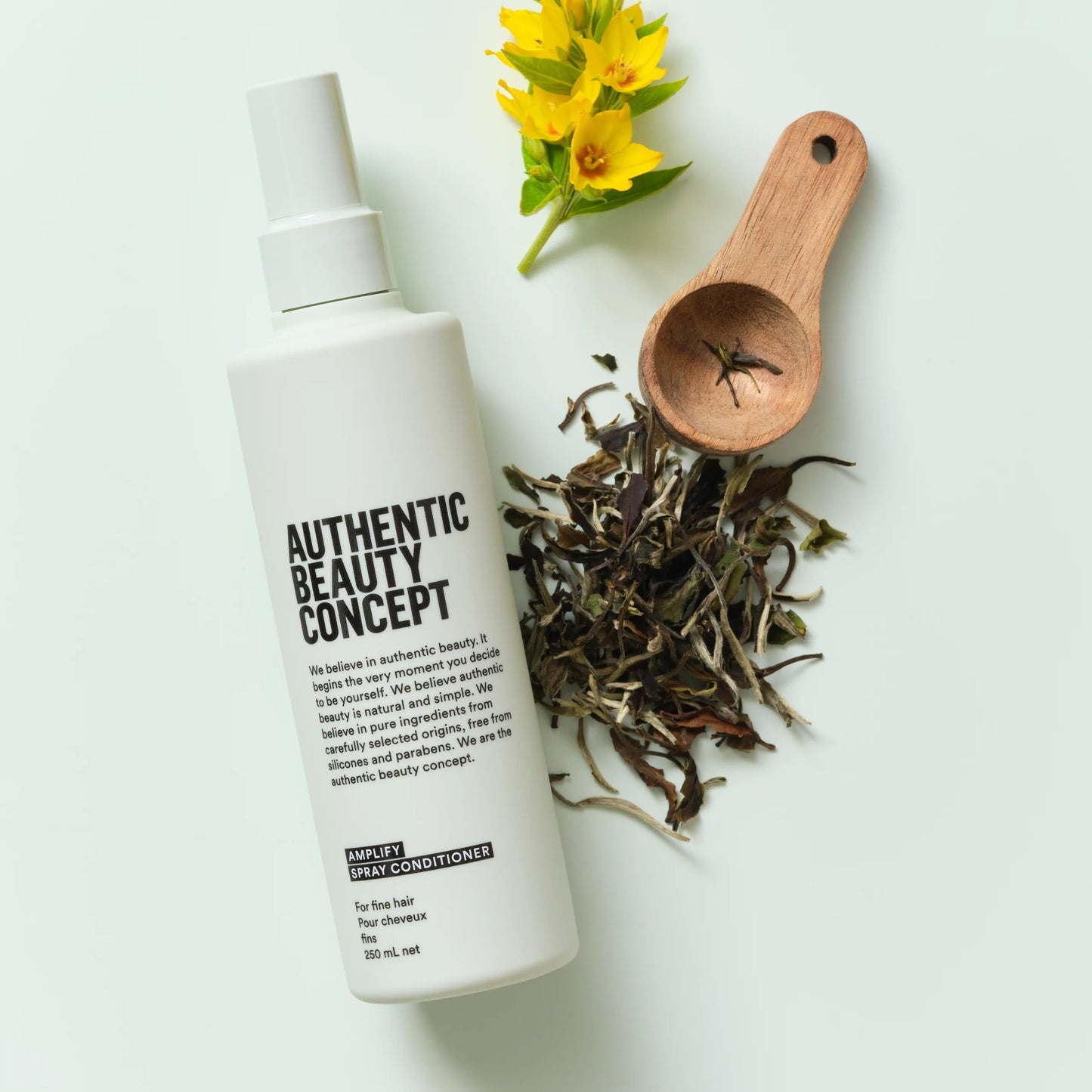 White bottle labeled 'Authentic Beauty Concept' with dried herbs, a wooden scoop, and yellow flowers on a light background.