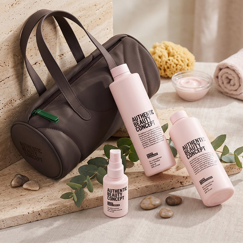 A dark brown barrel cosmetic bag with glow products and a glow serumfrom Authentic Beauty Concept, styled on a stone surface with eucalyptus leaves, pebbles, a sponge, and a rolled towel.