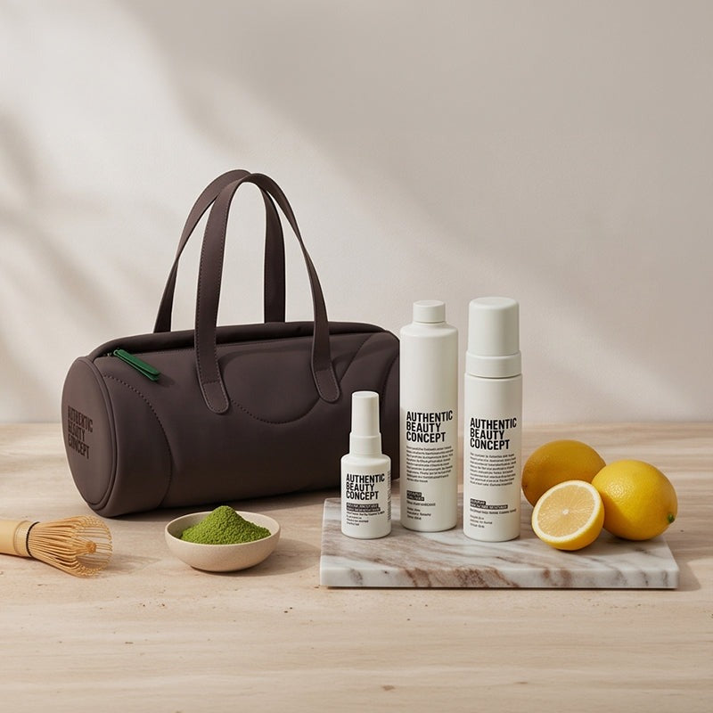A dark brown makeup bag with dual handles sits on a light wood table next to a marble tray holding three white cosmetic bottles from Authentic Beauty Concept, along with lemons, a small bowl of green powder, and a bamboo whisk.