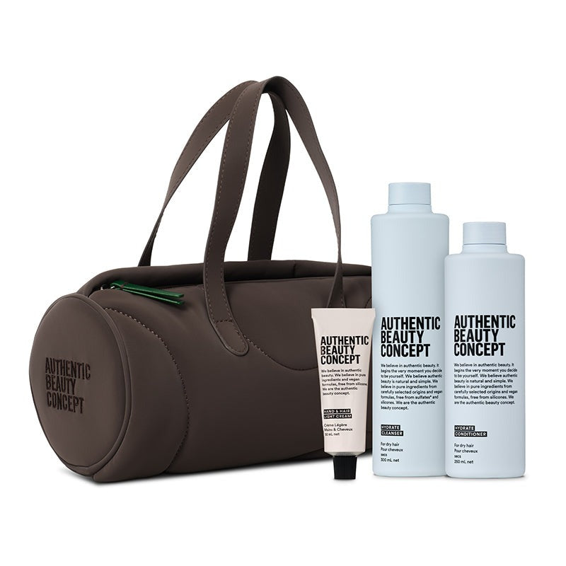A barrel brown cosmetic bag with two handles displayed alongside bottles of hydrate cleanser and conditioner and one small hand cream from Authentic Beauty Concept on a clean white background.