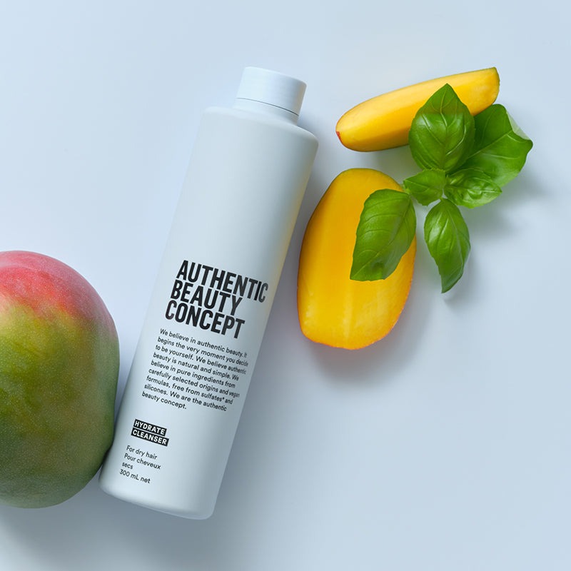 Hydrate Cleanser labeled 'AUTHENTIC BEAUTY CONCEPT'  is arranged on a light gray surface with a mango, a yellow mango slice, and green basil leaves as styling props.