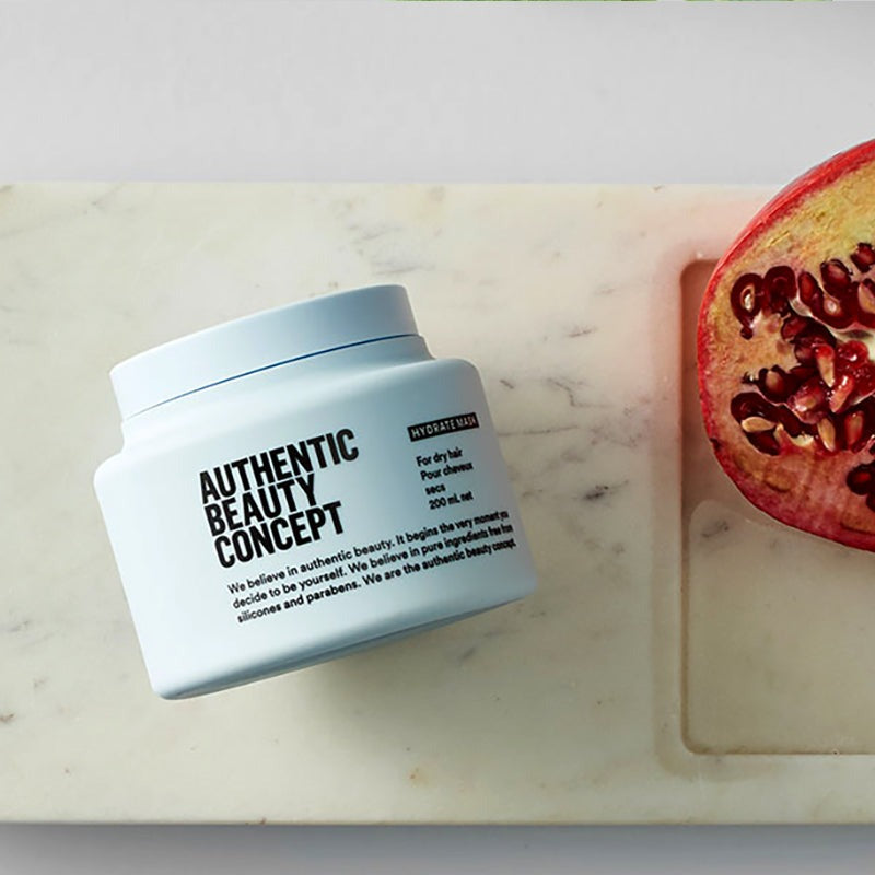 Hydrate mask labeled 'AUTHENTIC BEAUTY CONCEPT' placed on a light marble surface beside a sliced pomegranate half.