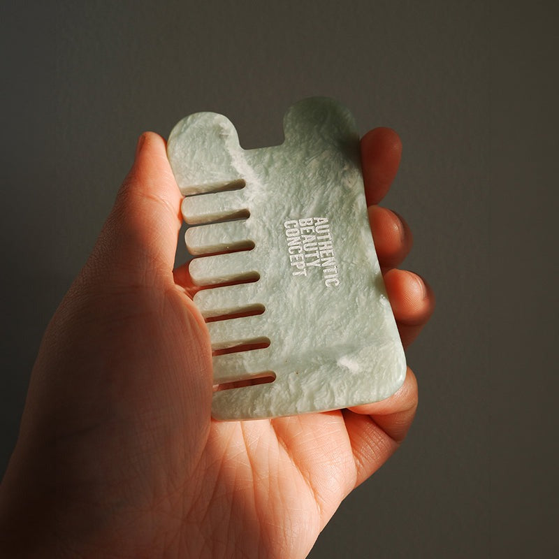 A hand holding a light green jade massage comb with the text 'AUTHENTIC BEAUTY CONCEPT' printed on it, set against a dark gray background.