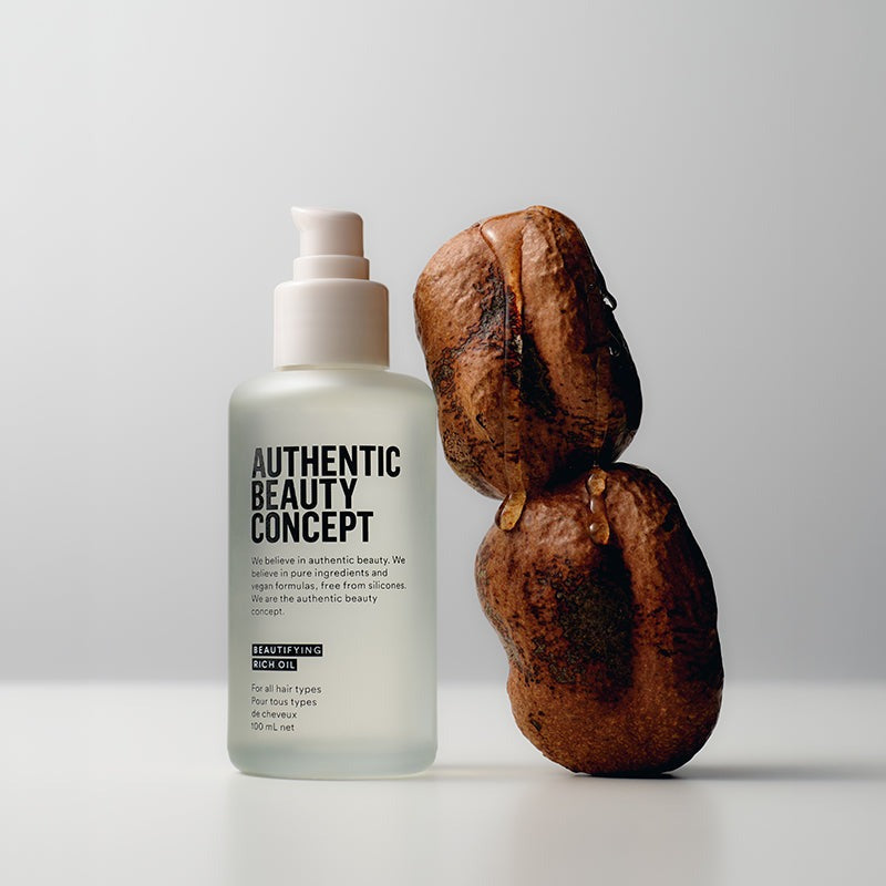Hair oil labeled 'Authentic Beauty Concept' next to a dried coconut on a light background