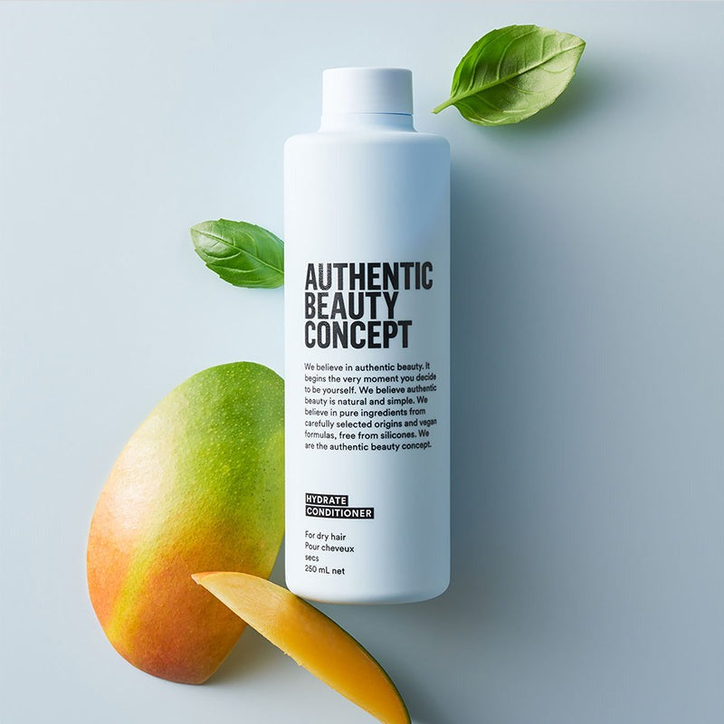 Hydrate cleanser of 'Authentic Beauty Concept' hair conditioner is styled on a light blue-gray background with a mango, a mango slice, and green leaves.