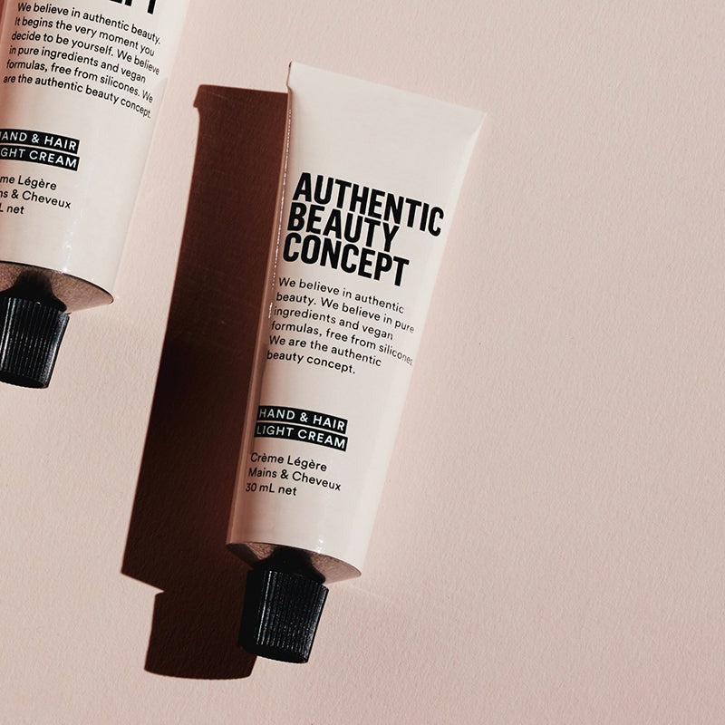 A squeeze tube of hand and hair light cream with black text on a white tube, set against a warm beige background with a soft shadow.