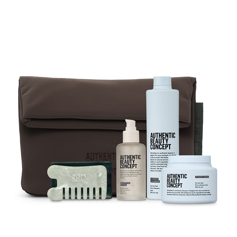 A hair care set from Authentic Beauty Concept, including a brown nylon pouch, a white bottle, a frosted pump bottle, a white jar, and a green-and-white massage comb, arranged on a white background.