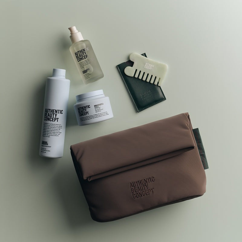 A flat-lay arrangement of skincare and haircare products on a light gray background, featuring a brown zippered pouch with 'AUTHENTIC BEAUTY CONCEPT' printed on it, alongside a white bottle, a white jar, a clear spray bottle, a cream comb, and a black cardholder.